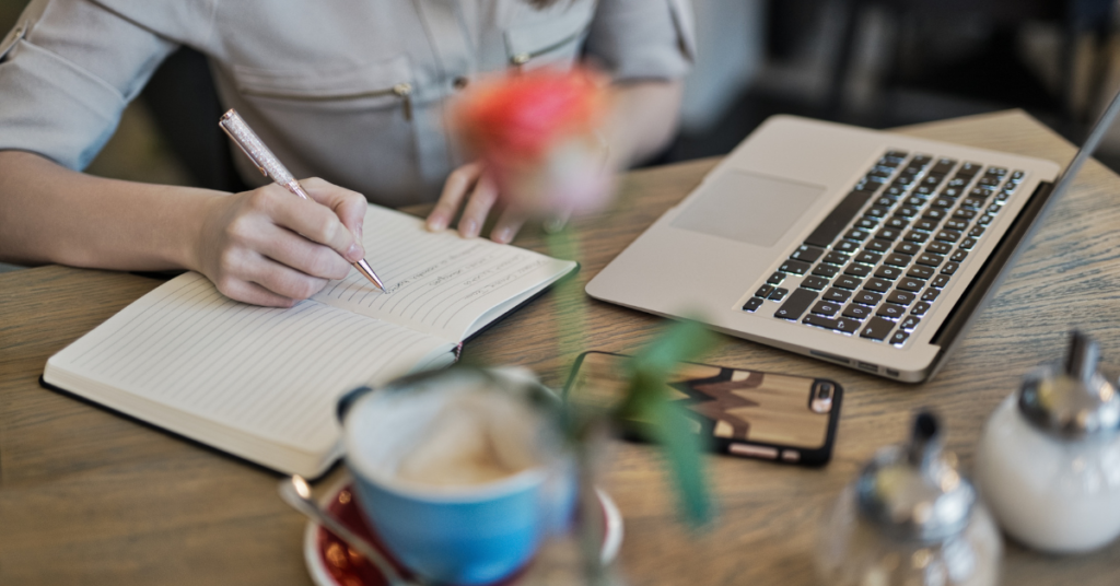 Close-up of a person's hands writing in a notebook, surrounded by tech and coffee, symbolizing productivity.